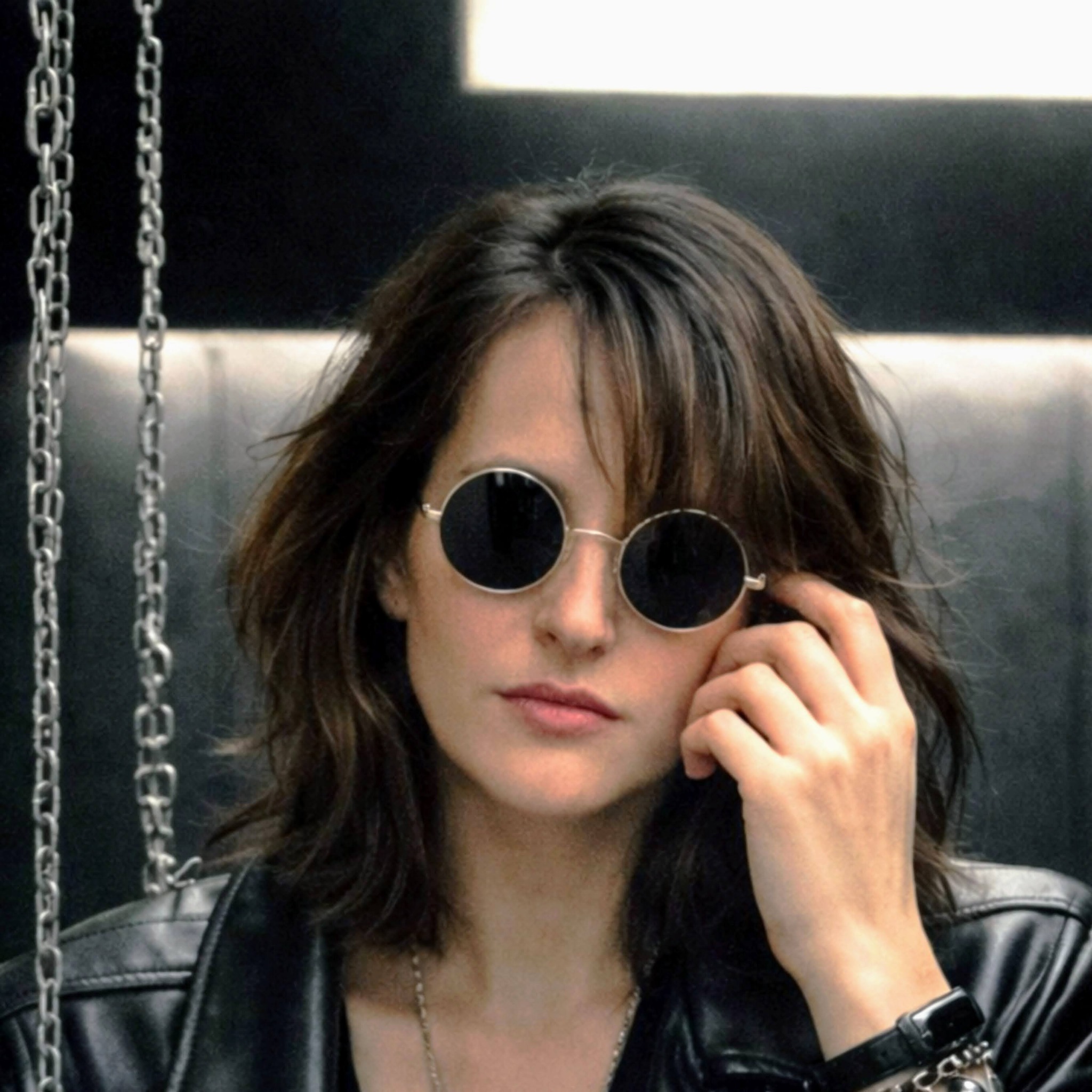 Madame Mystère en veste de cuir, la main près de ses lunettes rondes